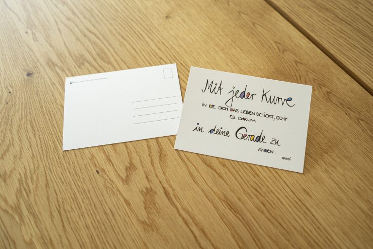 weiße Postkarte mit bunter Schrift "Mit jeder Kurve, in die dich das Leben schickt, geht es darum, in deine Gerade zu finden."
