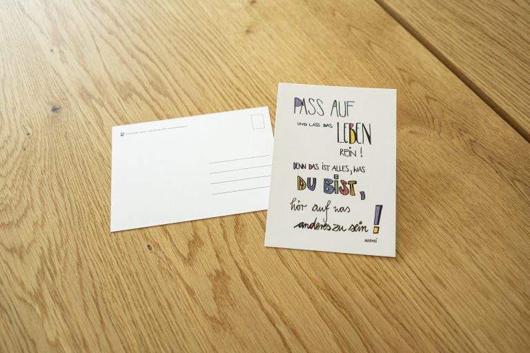 weiße Postkarte mit bunter Schrift "Pass auf und lass das Leben rein! Denn das ist alles, du bist, hör auf, was anderes zu sein!"